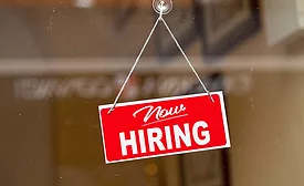 Red sign hanging at the glass door of a shop saying: "Now hiring".