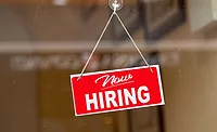 Red sign hanging at the glass door of a shop saying: "Now hiring".