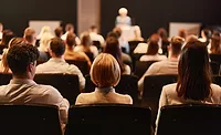 POV from a seat amongst an audience, listening to a speaker. Front rows and speaker are blurred.