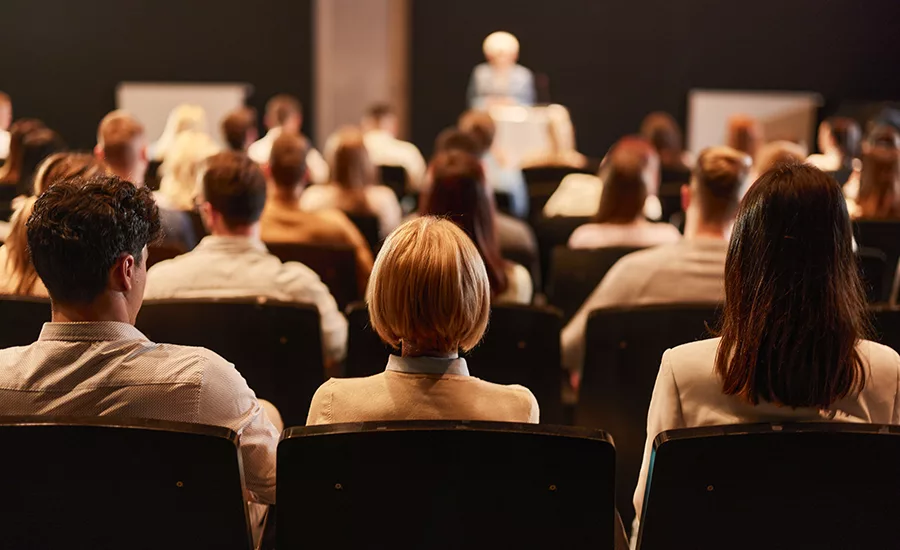 POV from a seat amongst an audience, listening to a speaker. Front rows and speaker are blurred.