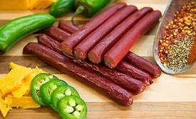 Dried meat sticks on a chopping board, whole and sliced jalapeno peppers, cheese and pepper seasoning on a board.