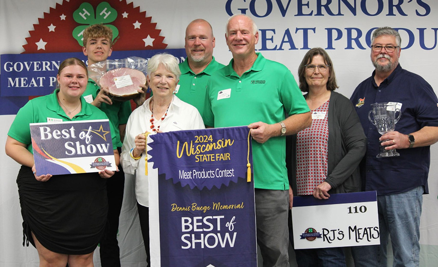 Winners posing for a photo at the Wisconsin State Fair meat contest, holding "Best of Show" signs, awards, ham, and trophy.