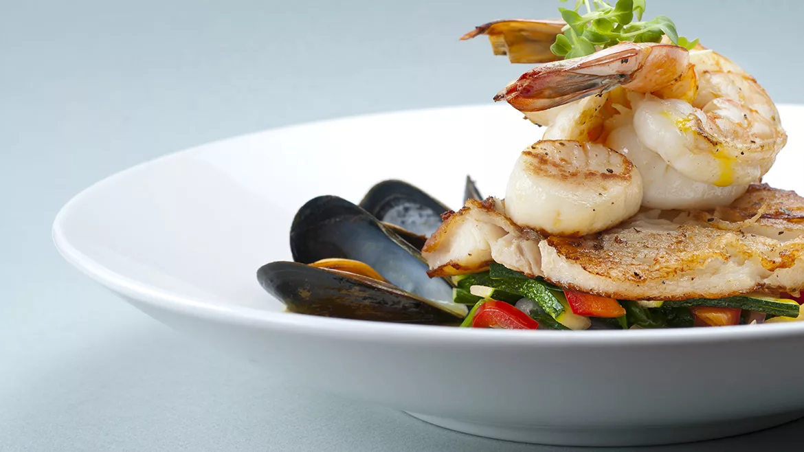 A white bowl of shrimp, scallops and muscles with a gray blue background.
