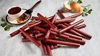 A stack of Lakeside Foods snack sticks on a table, a small cup of barbecue sauce on a small plate with a basting brush, and a cut onion with a coriander leaf above it.