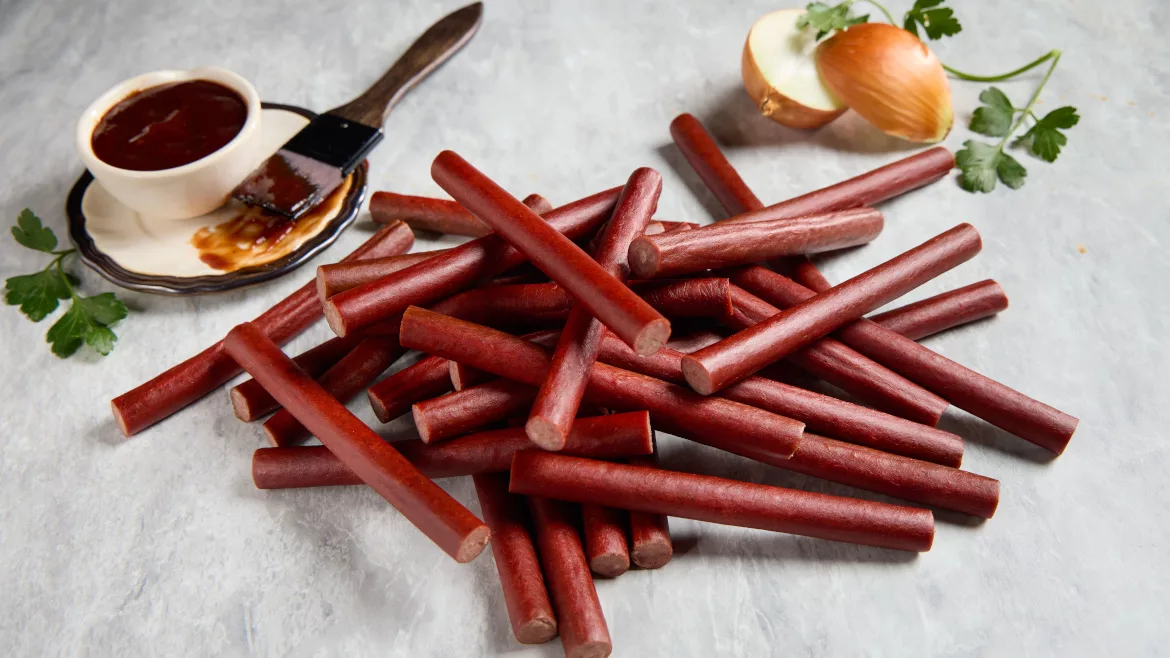 A stack of Lakeside Foods snack sticks on a table, a small cup of barbecue sauce on a small plate with a basting brush, and a cut onion with a coriander leaf above it.