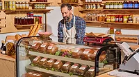 A shopkeeper in an apron stands behind a deli counter displaying packaged bread, with shelves of jars and condiments lining the walls.