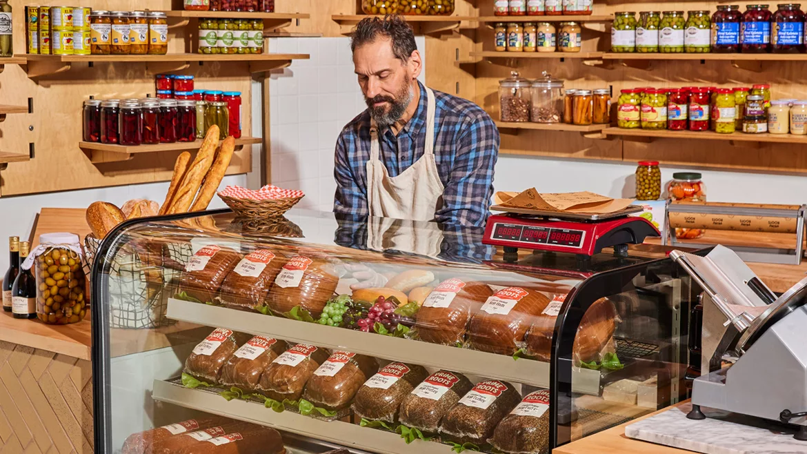 A shopkeeper in an apron stands behind a deli counter displaying packaged bread, with shelves of jars and condiments lining the walls.