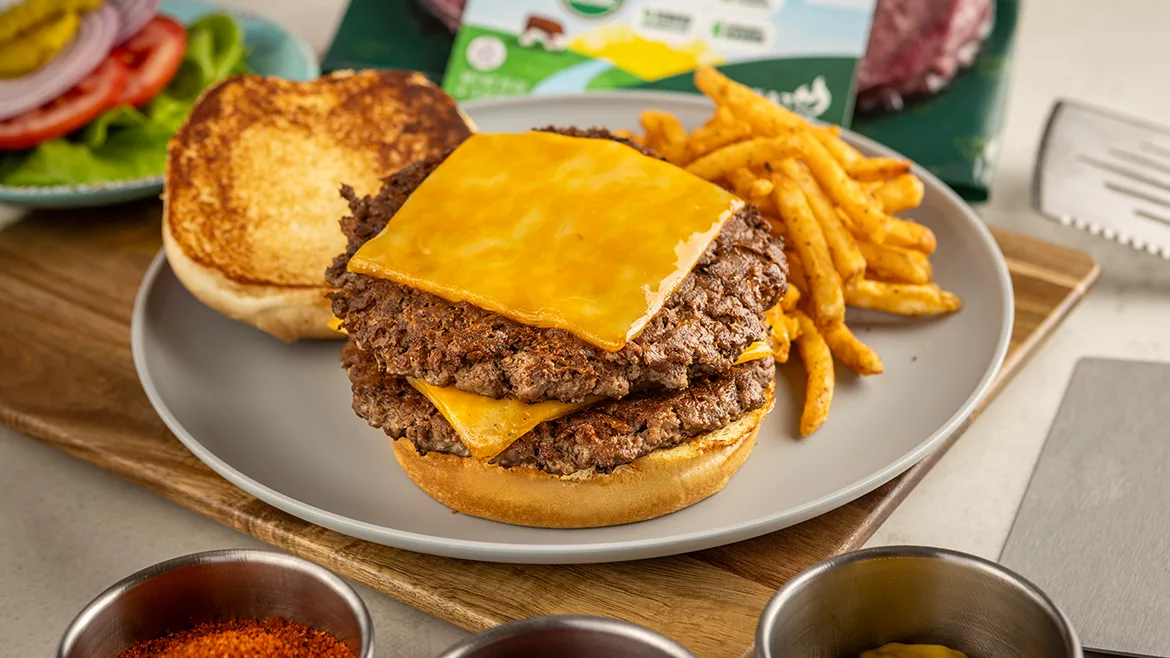 An open double cheeseburger on a white plate, the top bun beside it, along with a side of seasoned fries.