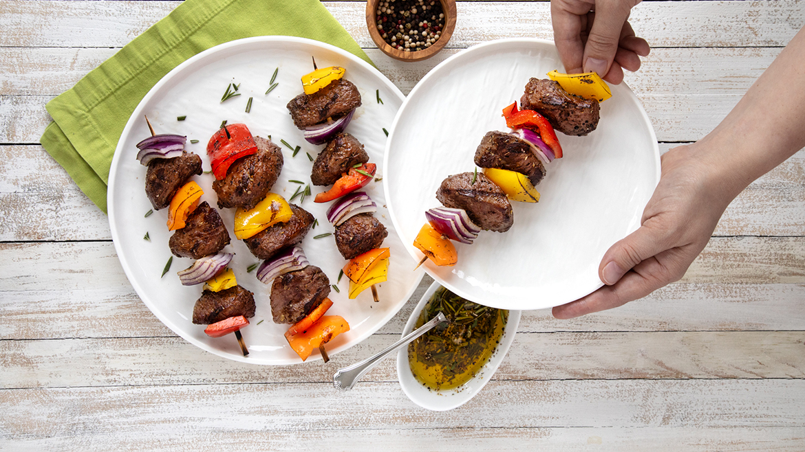Verde Farms Stew Meat on skewers with red onions red and yellow bell peppers on white plates. Small bowls of seasonings and a seasoned oil are also on the table.