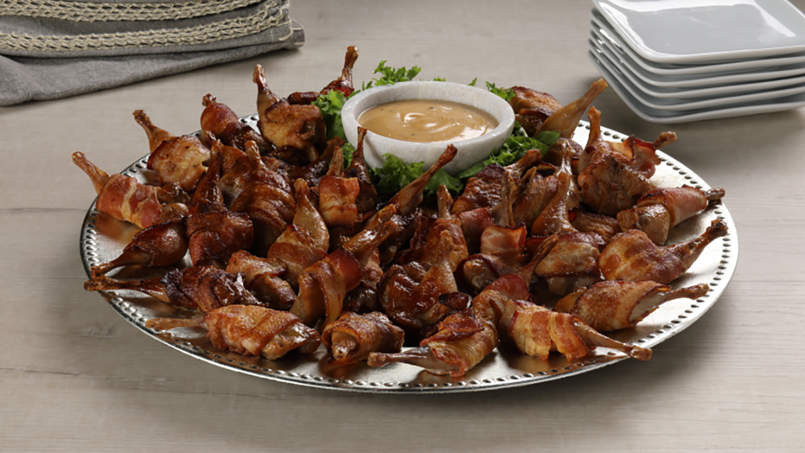 Bacon Wrapped Quail legs on a serving plate served with a dip.