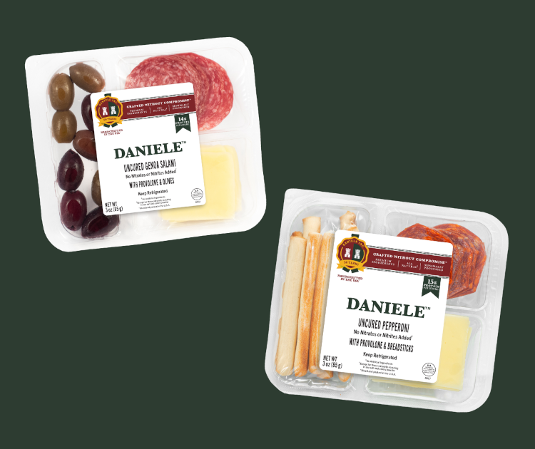 Two Daniele snack packs, one with olives, cheese and salami slices and the other with bread sticks, cheese and pepperoni slices on a dark green background.