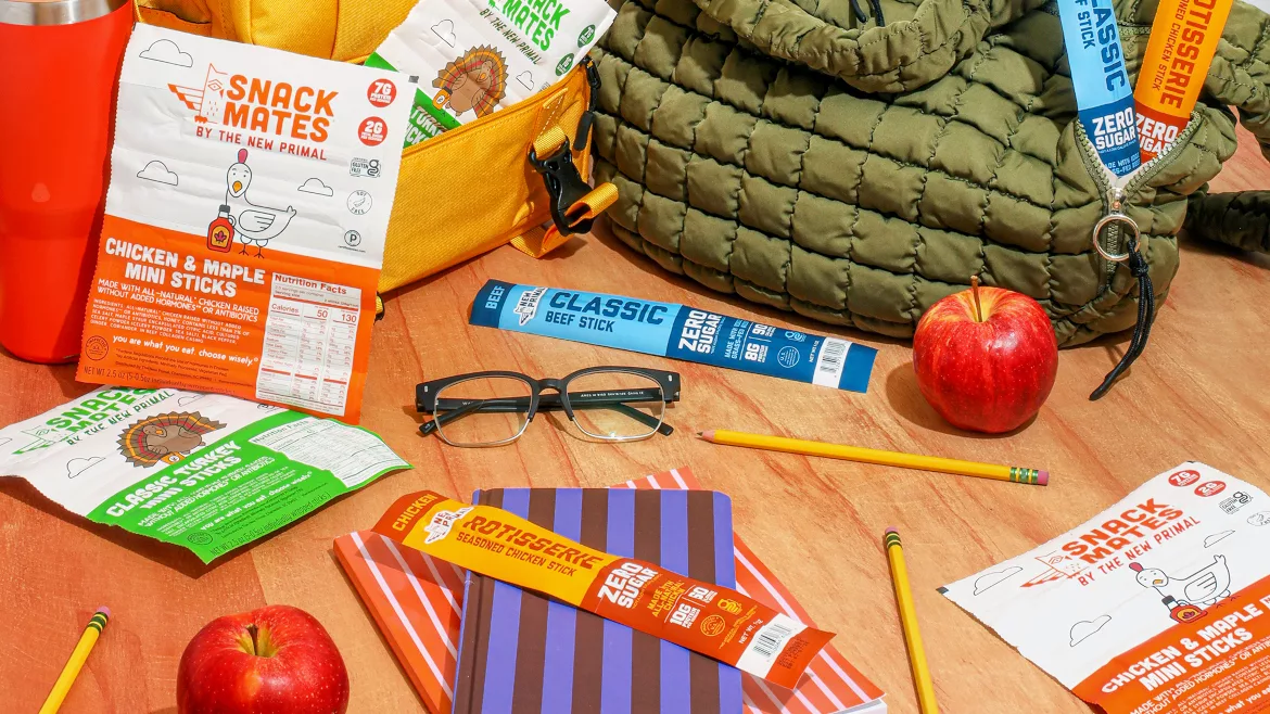Various new Primal snack sticks on a table amongst pencils, apples, a pair of glasses, lunch bags and a water bottle.