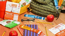 Various new Primal snack sticks on a table amongst pencils, apples, a pair of glasses, lunch bags and a water bottle.