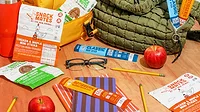Various new Primal snack sticks on a table amongst pencils, apples, a pair of glasses, lunch bags and a water bottle.
