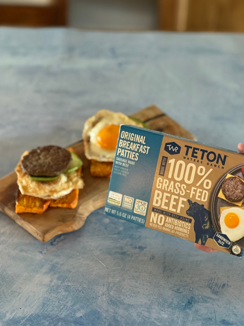 A hand holds a Teton Waters Ranch 100% Grass-Fed Beef Original Breakfast Patties box next to prepared sandwiches.