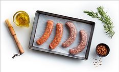 A cooking tray of raw sausages lined with parchment, a cup of olive oil herb mixture, wooden basting brush, a small bowl of seasoning and a sprig of rosemary on a white table.