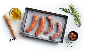 A cooking tray of raw sausages lined with parchment, a cup of olive oil herb mixture, wooden basting brush, a small bowl of seasoning and a sprig of rosemary on a white table.