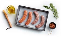 A cooking tray of raw sausages lined with parchment, a cup of olive oil herb mixture, wooden basting brush, a small bowl of seasoning and a sprig of rosemary on a white table.