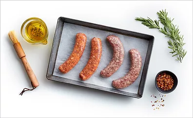 A cooking tray of raw sausages lined with parchment, a cup of olive oil herb mixture, wooden basting brush, a small bowl of seasoning and a sprig of rosemary on a white table.