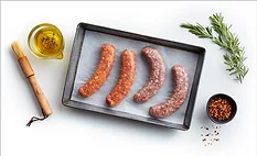 A cooking tray of raw sausages lined with parchment, a cup of olive oil herb mixture, wooden basting brush, a small bowl of seasoning and a sprig of rosemary on a white table.