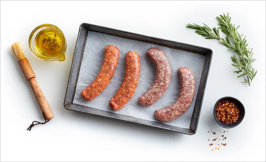 A cooking tray of raw sausages lined with parchment, a cup of olive oil herb mixture, wooden basting brush, a small bowl of seasoning and a sprig of rosemary on a white table.