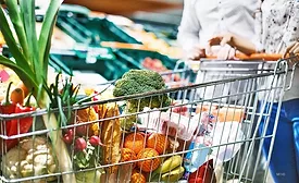 Close up of a grocery cart full of groceries, a cropped image of a couple pushing the cart and a blurred background of the vegetable aisle.
