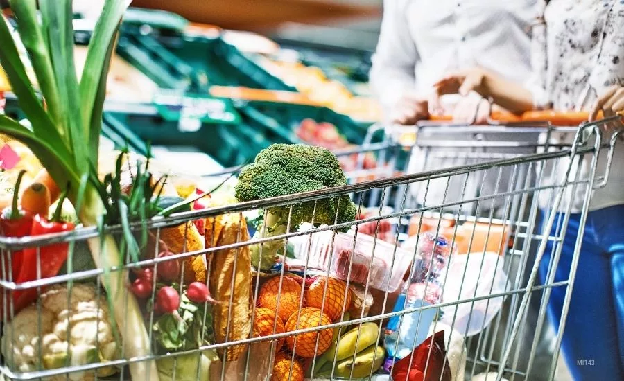 Close up of a grocery cart full of groceries, a cropped image of a couple pushing the cart and a blurred background of the vegetable aisle.