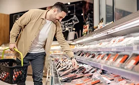 A smiling man carrying a grocery basket is reaching down to pick up a package of meat in a grocery store.