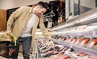 A smiling man carrying a grocery basket is reaching down to pick up a package of meat in a grocery store.