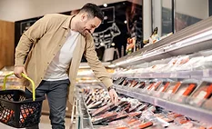 A smiling man carrying a grocery basket is reaching down to pick up a package of meat in a grocery store.