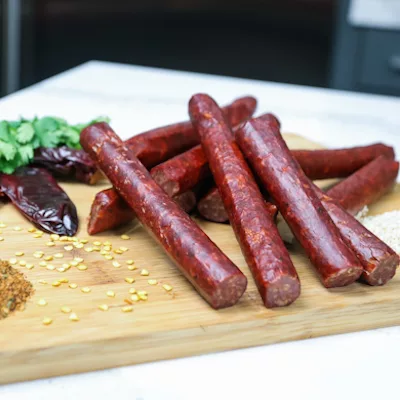 Seasoned sausages stacked on a wooden board along with various seeds, peppers and herbs.